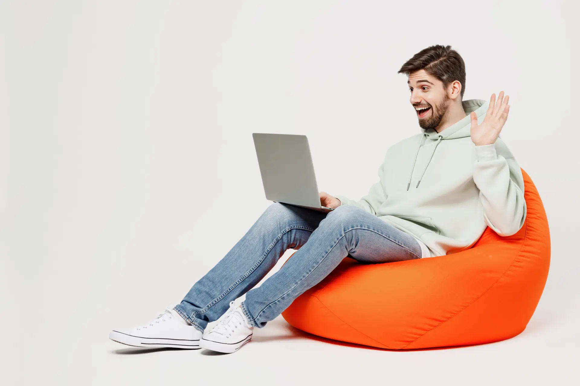 Full body young smiling fun IT man wears mint hoody sit in bag chair hold use work on laptop pc computer waving hand get video call isolated on plain solid white background People lifestyle concept